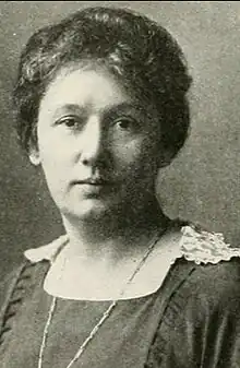 A white woman with hair dressed to the nape, wearing a dark dress with a square neckline and a lace-trimmed collar