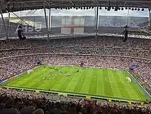 The interior of an empty stadium as viewed from its upper tier of seating. The seats are a vivid red and the pitch is a vivid green. The pale grey sky is visible through an opening in the ceiling above the pitch.