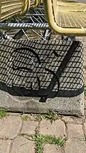 The bench consists of a metal yellow square grid pattern seating surface supported by a tubular steel girder truss in between two concrete blocks with the Expo 86 logo debossed on the outer side of the block.