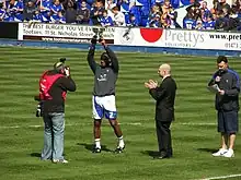 Four men standing on a grass football pitch. The man second from left, wearing a grey top, white shorts and white socks, is holding a trophy above his head. Spectators wearing blue or black tops are visible in the background.