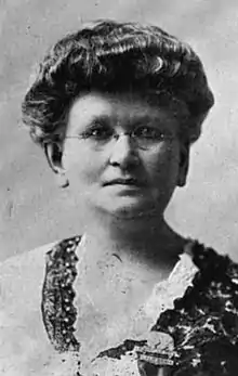 A white woman with thick wavy hair in a bouffant updo. She is wearing a lace collar and eyeglasses.