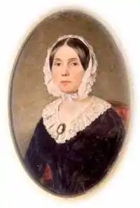 Oval of young woman seated, with pinkish white frilled head bonnet and dress top, black narrow waist dress, straight dark hair parted in the middle