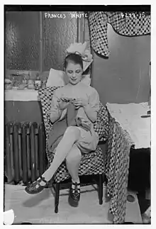 A young white woman wearing a theatrical costume, seated indoors, knitting