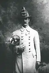 A man in a white coat and fire helmet holding a brass horn