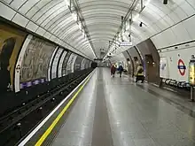 The interior of a building with a rounded roof and walls, several doors on the right, railway tracks on the left, and three people walking in the middle