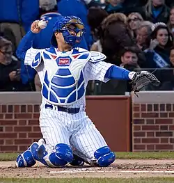 A man kneeling in a white baseball uniform with blue pinstripes and catcher's gear