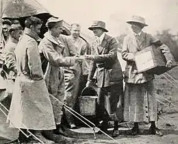 A group of men approach two women, all in uniform; one of the women is holding a large box. They are oundoors and there are tent ropes visible in the frame.