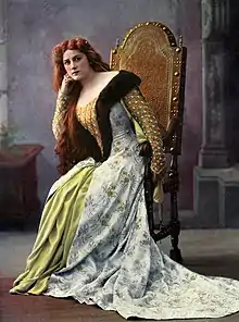 A white woman, seated, wearing a costume and long red hair, as Marguerite de Bourgogne. The image is hand-colored.