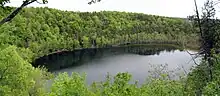 A small lake seen from the top of the cliffs surrounding it. There are trees growing on the steep slopes; their leaves are light green.