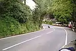 Racing motorcycle negotiating the entrance to Glen Helen turn, exiting uphill, set deep in the countryside with the entrance to the actual Glen and car park (shared with the closed restaurant) to the right, with many spectators around the outside of the bend