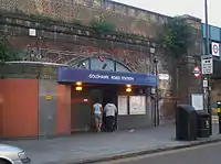 A brown-bricked building with a blue sign reading "GOLDHAWK ROAD STATION" in white letters and two women walking in front all under a grey sky