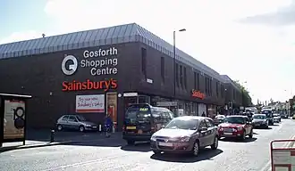 A 1970s monolithic shopping centre, mostly grey/brown in colour. In the foreground the busy high street with a number of travelling cars.