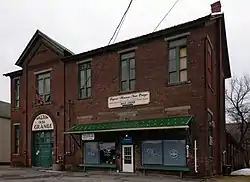 A dark red brick two-story building under an overcast sky, seem from slightly to its left. There is a chimney rising from its right end, and a projecting gabled portico on its left. At right on the ground floor is a hair salon, and at left the transom over a large door in the portico says "Walton 1454 Grange".