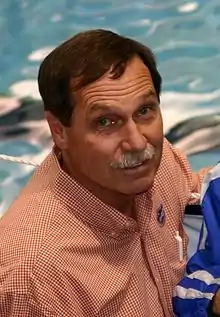 Head and shoulders portrait of Gregg Troy, dark-haired, 58-year-old man with grey mustache, shown in an orange-and white checkered shirt.