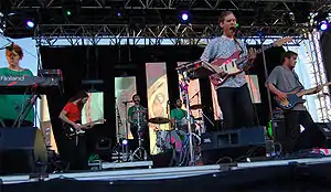 A six member group is on stage. At the left a woman is standing behind keyboards, further back is a man with a long beard playing guitar and he is half turned in right profile. At the back of the stage is a man singing and playing an instrument, beside him a seated man with a short beard is playing drums. At stage front is a man singing and playing guitar and then a man in right profile playing a guitar.