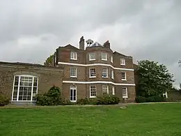 A three-storey brown brick building with a cupola, and a single storey extension on the left, the foreground is a green lawn