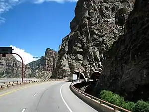 Under the clear sky, the freeway enters a tunnel through a mountain which is covered by solid rock.