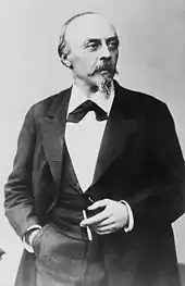 A middle-aged, balding man with a moustache and small beard, wearing a dark suit and holding a cigarette.