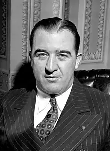 A man with slicked-back black hair wearing a pinstriped suit, patterned tie, and white shirt