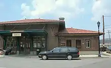 Entrance to a train station, signs read "Hastings-on-Hudson", "Station Cafe", "MTA Metro-North Railroad". Two people are sitting out front drinking coffee, and a black Mercedes wagon is parked.