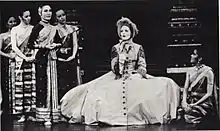 Black-and-white photo of a stage scene.  A Caucasian woman wearing a bonnet sits on a chair at center, with her huge Victorian hoop skirt spread out around her, singing.  Several women in elegant silky Asian clothing surround her.  One at right (Tuptim) kneels watching her, as the others stand in a clump at left.