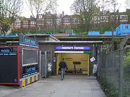 A brown-bricked building with a rectangular, blue sign reading "HIGHGATE STATION" in white letters and a person walking in all under a white sky