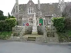 Stone cross, with red wreaths, separated from a building behind by metal railings.