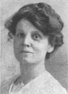 A white woman with dark hair in a bouffant updo, wearing a white lace-trimmed blouse