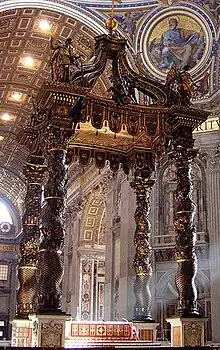  Photo shows the baldachin standing in the centre of the church, viewed looking towards the nave. There is an altar beneath it which has a red and gold frontal cloth decorated with large crosses.