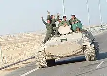 Six uniformed soldiers waving from an armoured vehicle on a highway