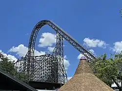 Appearing from the central plaza, Iron Gwazi's lift hill is prominently shown in the skyline with the barrel roll-down drop under the lift structure. Parts of the building that houses the hybrid roller coaster are imaged at the bottom, with a thatched roof in the center-right. Trees are also visible on either side of the image.