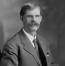 A man with dark hair and a mustache wearing a dark jacket, patterned tie, and white shirt