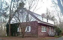 A house built of red stone house with a curved, sloping roof and white wooden top, seen from the side. It has brick chimneys at either end and some ivy on the visible side.