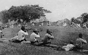 A dozen prisoners sit bound at a distance as Japanese soldiers in the foreground kill them