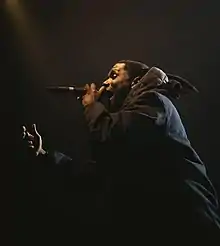 A black man, dressed in all black, stands side on to the camera. He is performing on stage with a microphone in his hand