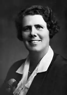 A smiling white woman with short wavy dark hair, wearing a white blouse with a sprad collar, over a dark jacket or cardigan