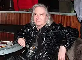 A color photograph of a seated man, with his right arm on a table, looking toward the camera.