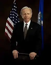 A man in a formal suit standing in front of national and state flags.