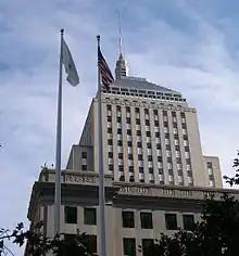 Ground-level view of an Art Deco-style skyscraper with a tan facade; the building is adorned with a thick, gray spire. In the foreground, two flags are visible.