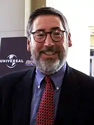 Headshot of a middle aged. Looking and smiling directly into the camera, the man wears rounded spectacles and sports a light grey beard. He wears a suit jacket, with a blue shirt and a patterned tie.