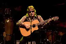 A man sitting, holding a guitar, with a microphone in front of him.