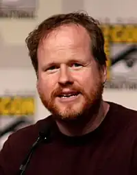 A bearded man, wearing a garnet shirt, speaking in front of a microphone. Behind the man is an out-of-focus white and yellow background.