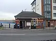 A red-bricked building with a rectangular, dark blue sign reading "KENTON STATION" in white letters all under a blue sky with white clouds