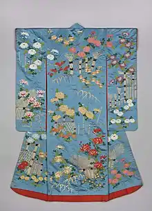 A light blue kimono viewed from the back. It has long sleeves, a bright red lining and padded red hem, and is decorated with bamboo bridges, fences and chrysanthemums in white, orange, yellow, red pink and gold.