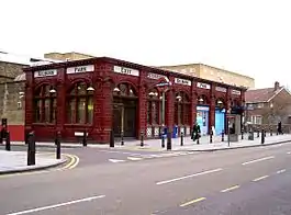 A red-bricked building with a series of white signs together reading "KILBURN PARK STATION" in red letters and people walking in front all under a white sky