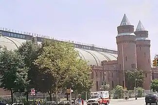 A large round-roofed building running the width of the image behind a city street and trees. At the right are two conical towers and an entrance pavilion