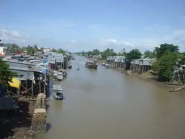 Brown-coloured waterway, with many small boats and elevated wooden and thatched houses visible. Foliage is also in abundance.