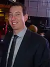 A clean shaven man in his early thirties wearing a black suit, a white shirt and a gray tie