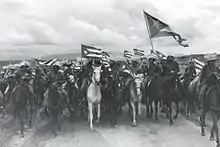 Black and white photo of riders seen from the front carrying the Cuban flag.