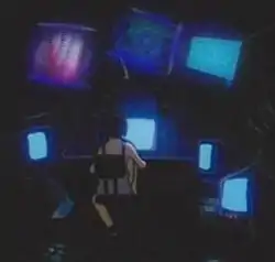 A young girl in a white shift sits with her back to us in the dark, focusing her attention on many glowing computer screens which surround her.